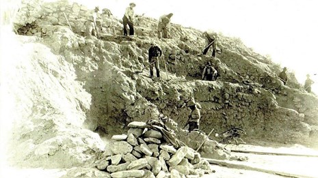 Sepia photo of seven people working to excavate and reconstruct stone walls