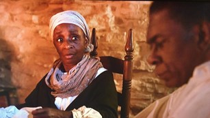 Screenshot of an African-American woman in period clothing, African-American man in the foreground.