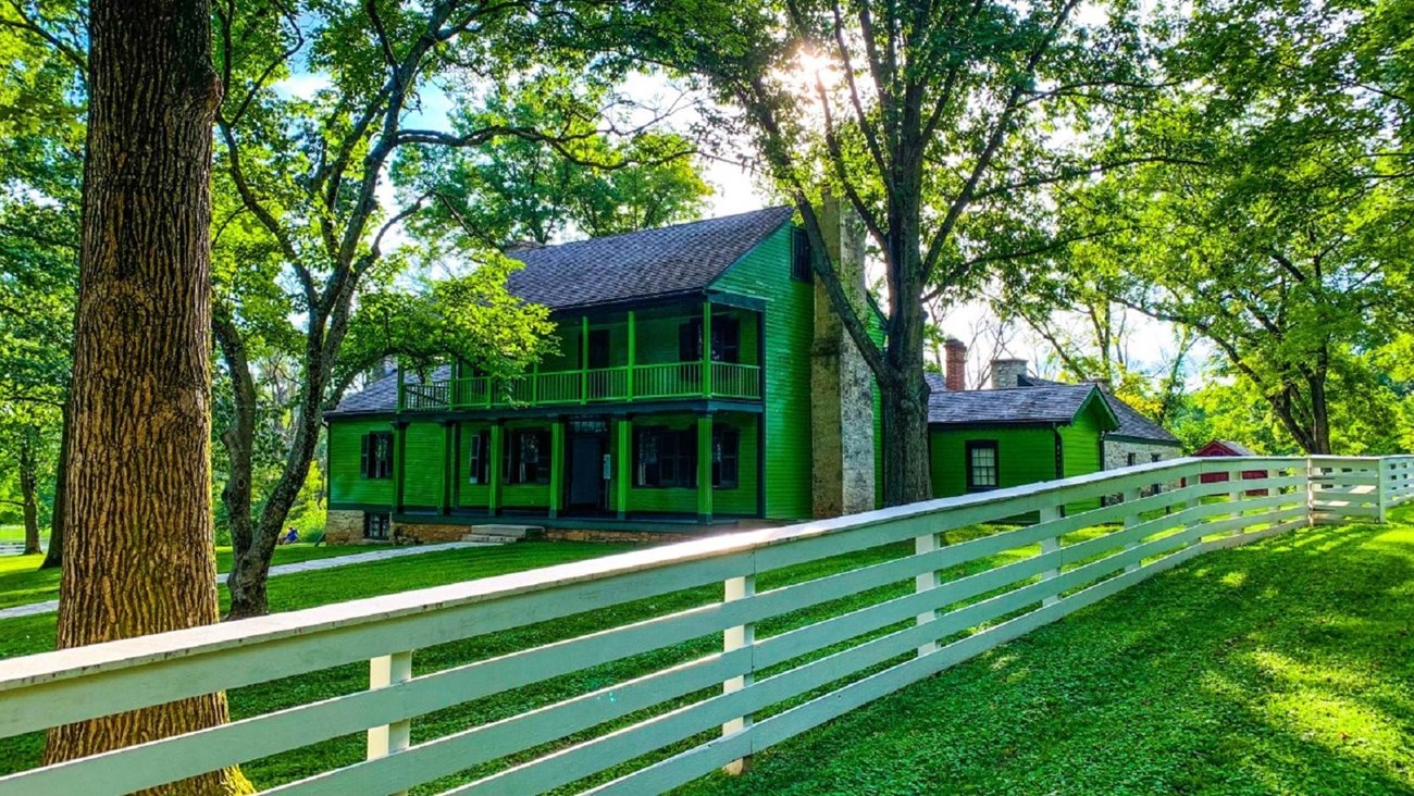 Green two-story frame house surrounded by trees and a white fence. 