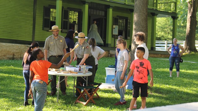 Two Park Rangers wearing green and gray uniforms interacting with five students in front of a house.