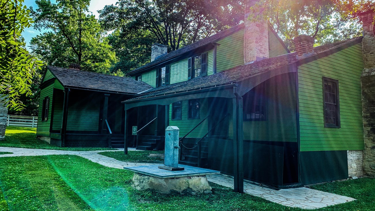 Green two-story frame house surrounded by trees with sunlight in background.