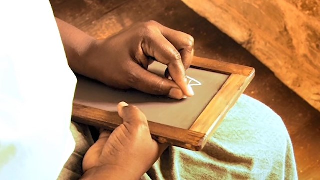 African American wearing a white dress writing alphabet on a chalk board.  