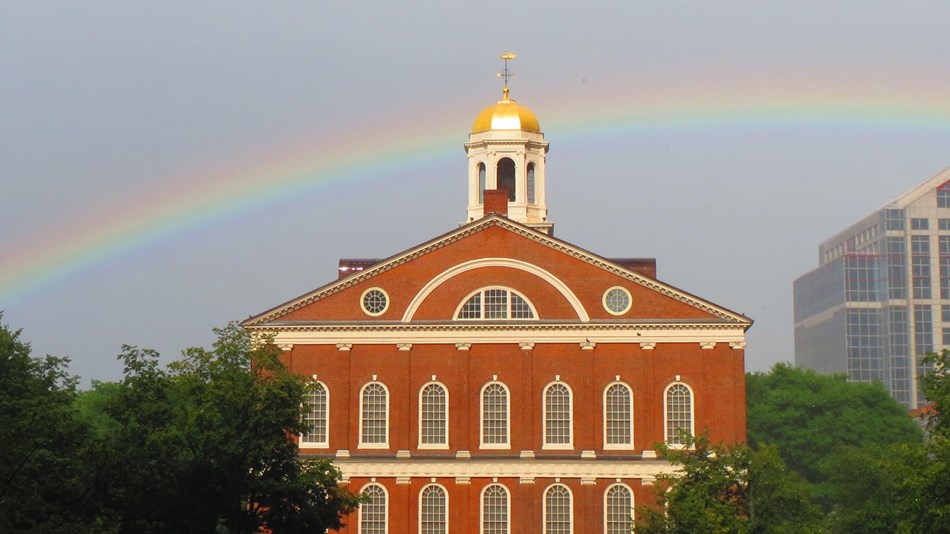 A rainbow stretches behind Faneuil Hall - a four story brick building with a gold domed cupola.