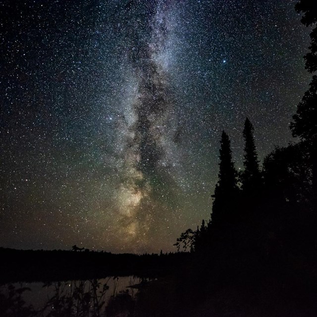 A photograph taken of the night sky with hundreds of stars above a lake and a forest.