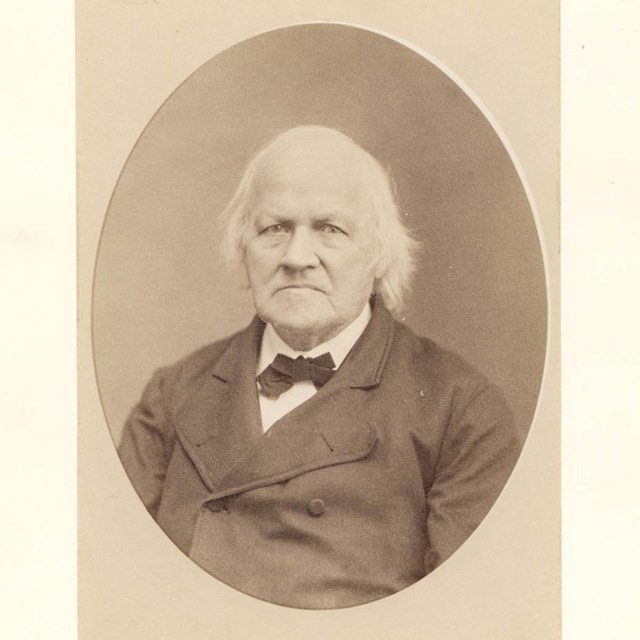 Photograph of a white man with shoulder length white hair in a suit.