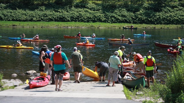 boat access ramp to river. Pairs of people in T-shirts and shorts carry kayaks and canoes to water.