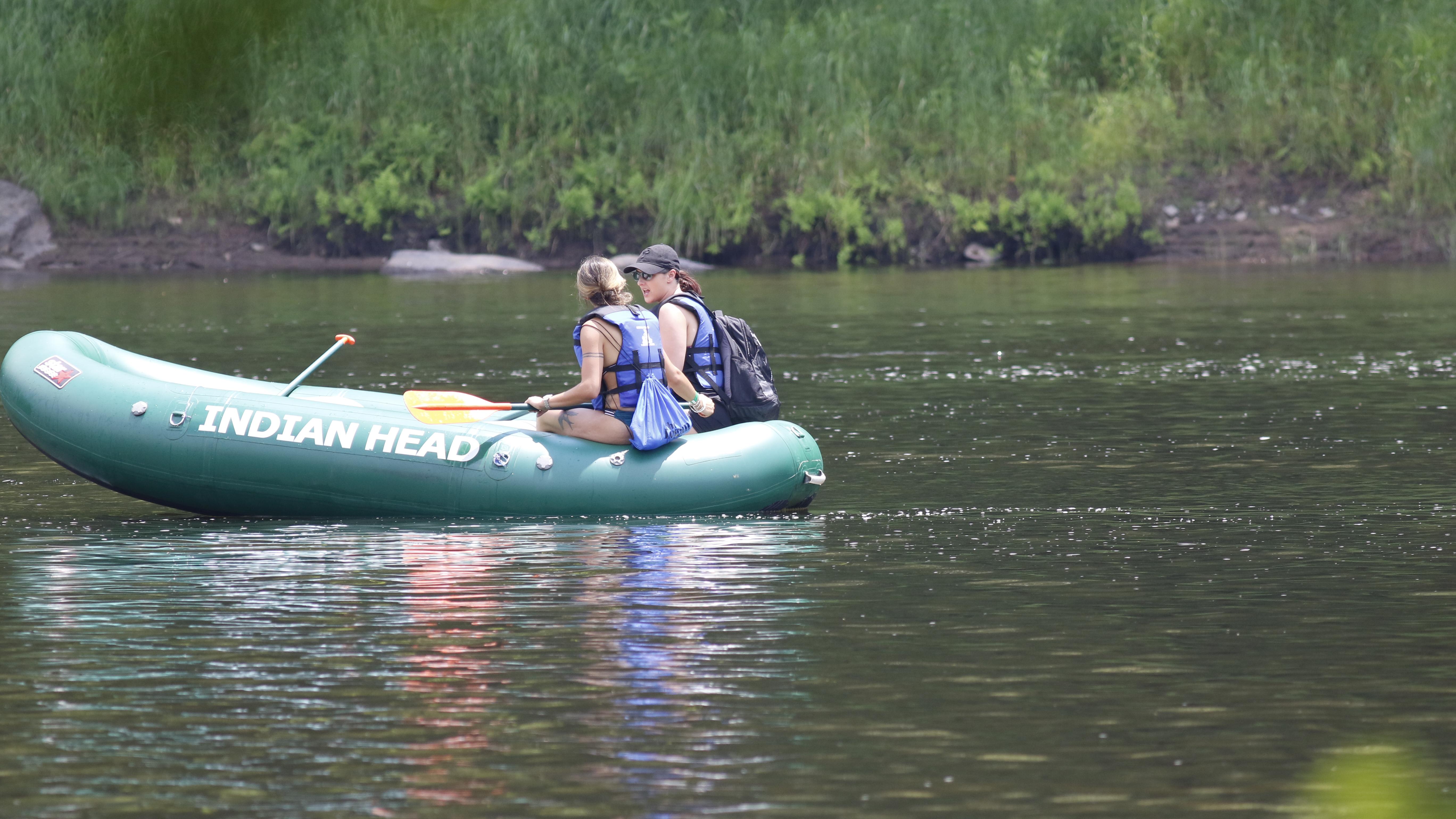 2 people with life jackets in green inflatable raft in river. Raft has printed text "Indian Head."