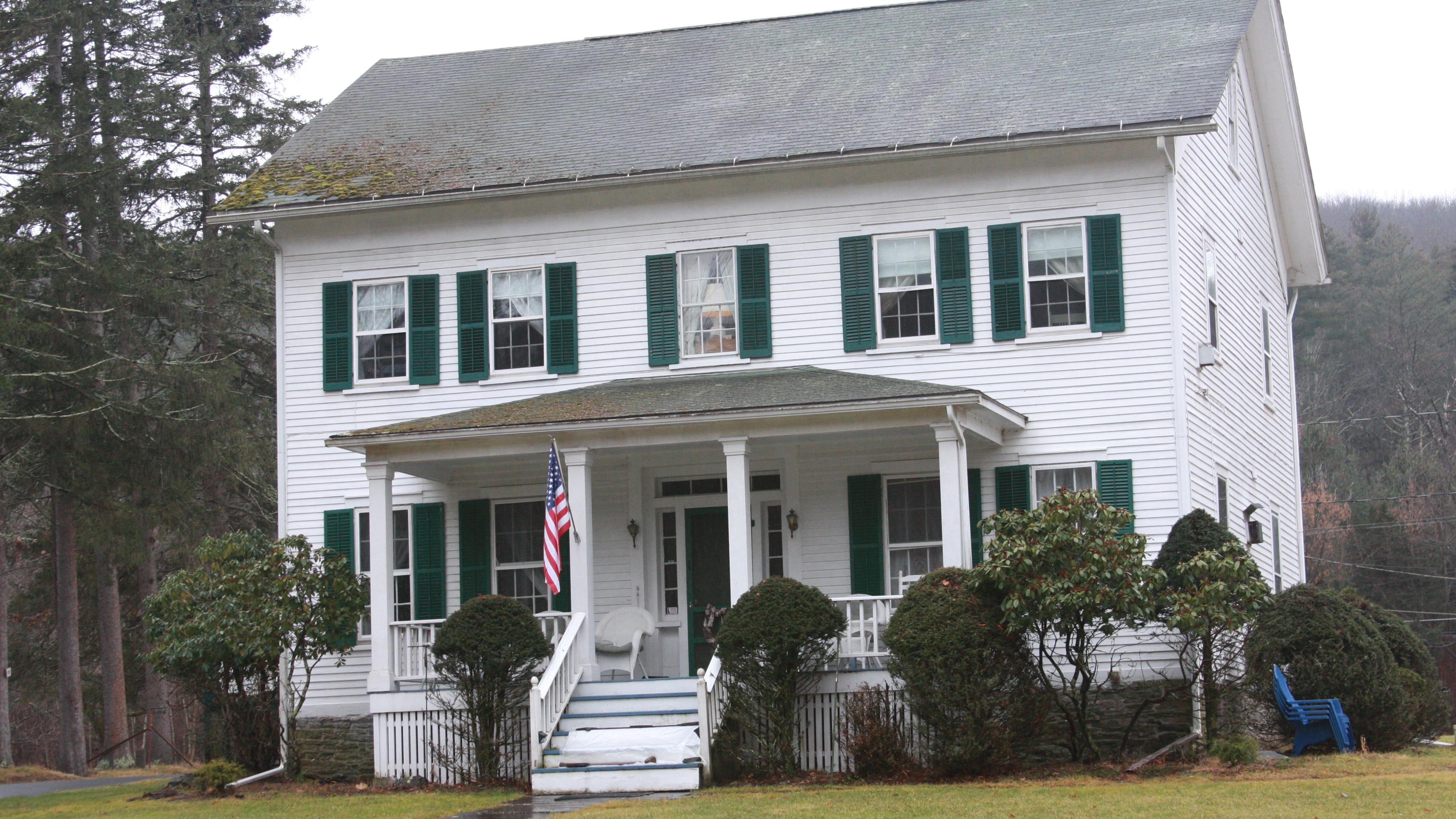 White two-story wood frame house with grey A-frame roof, dark green shutters, and small front porch 