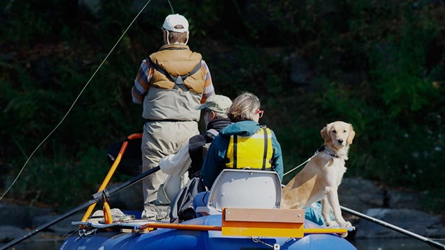 Two men, one woman, and dog sitting in inflatable raft with fishing and paddling equipment.