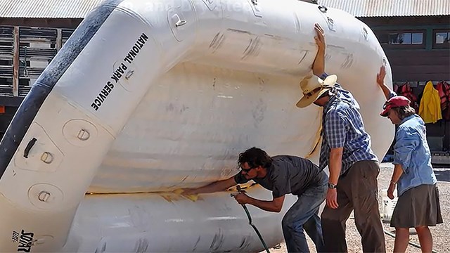 Two men hold a white raft upright on its side while another man sprays the inside with a hose