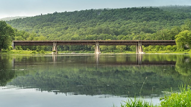 View of Roebling's Delaware Aqueduct over the Delaware River. Forested hill reflects on water.