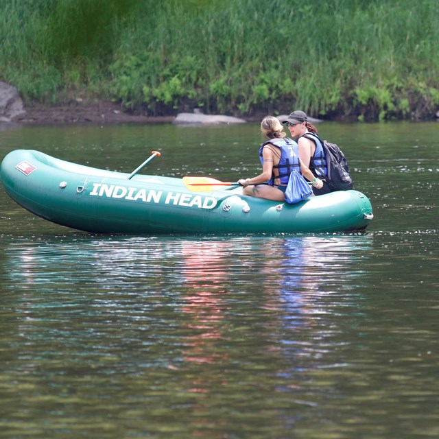 2 people with life jackets in green inflatable raft in river. Raft has printed text 