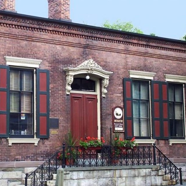 Low brick building with tall windows, blue and red shutters, and fancy trim.