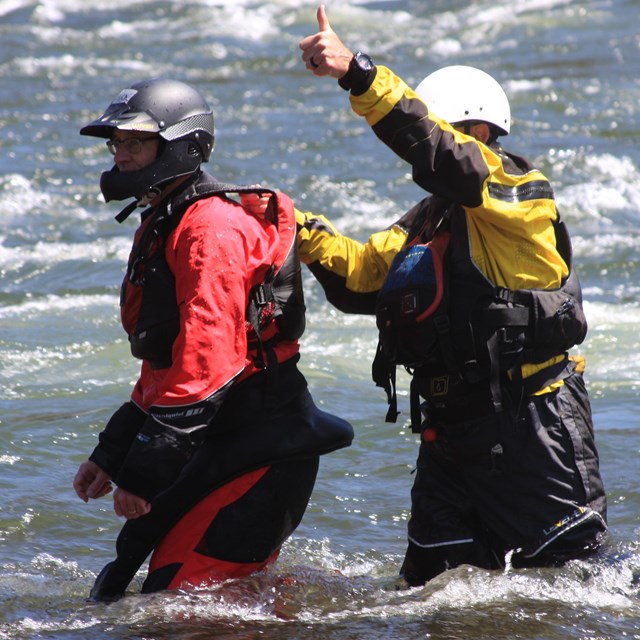 two people in helmets, lifejackets, and wet gear in river. One gives a thumbs up.