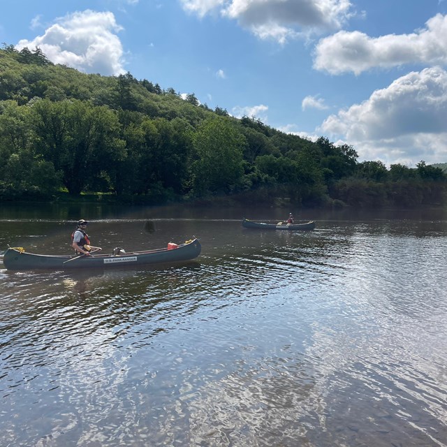 Rangers in canoes on the river. Blue sky with fluffy clouds. Green trees cover hills on river.