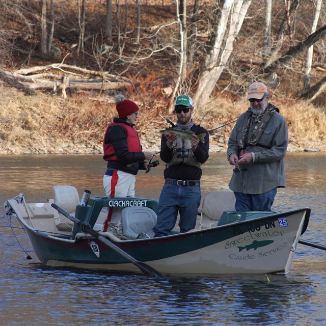 three people standing in boat with motor. Man in middle holds up large, green-gray fish.