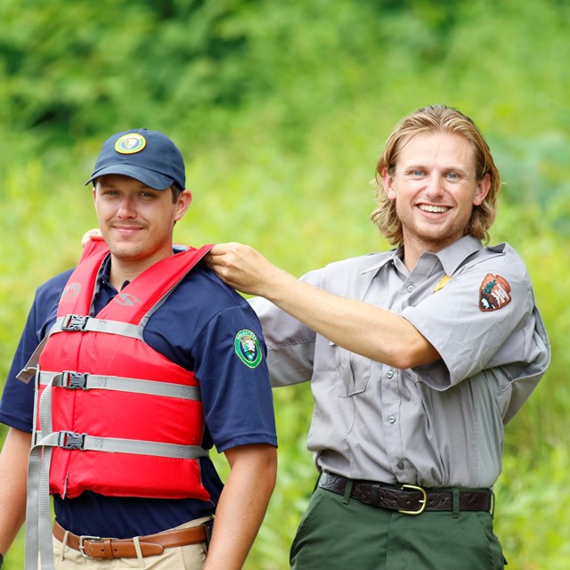Park Ranger tugs on life jacket worn by intern at shoulders. Both smile at camera.