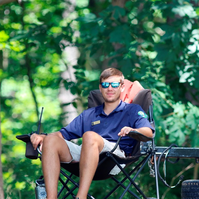 Young man in reflective sunglasses lounges in camping chair in forest.