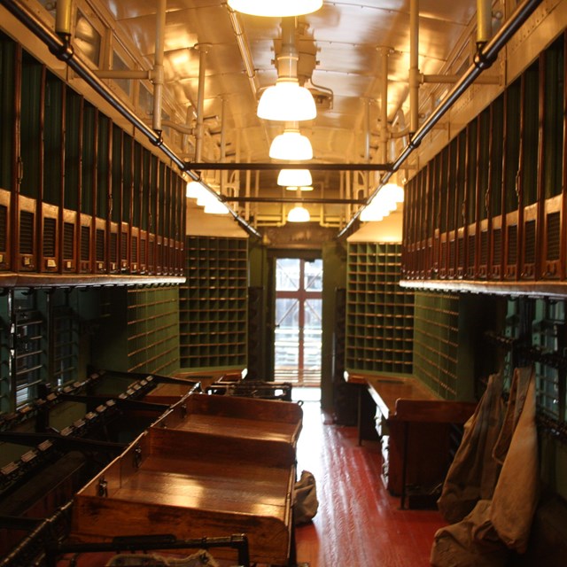 View down an old mailroom in soft yellow lamplight. Red and dark green mailboxes line walls.