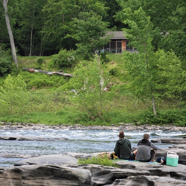 Couple sits on rocks on the edge of a river. Yellow-green trees form a forest across the river.