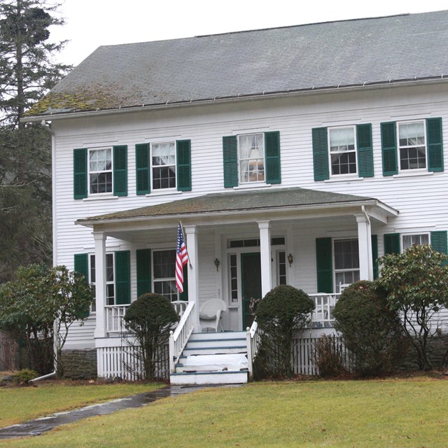 White two-story wood frame house with grey A-frame roof, dark green shutters, and small front porch 