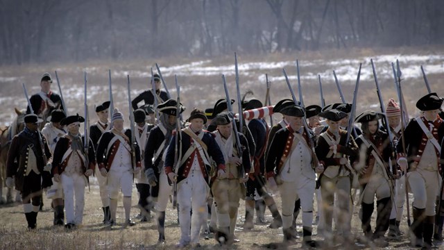 Tired-looking Revolutionary War soldiers hold muskets against their shoulders and march through mud