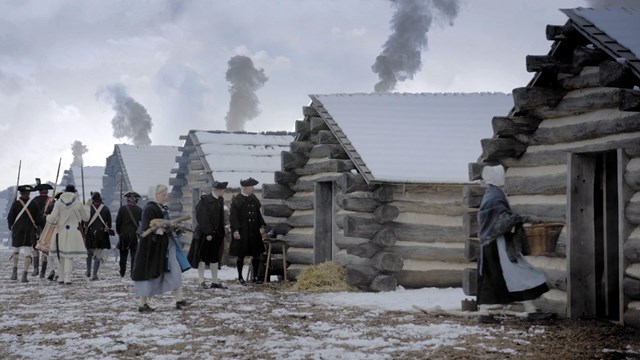 Soldiers in uniform and women wearing cloaks walk in front of a line of snow-covered log huts.