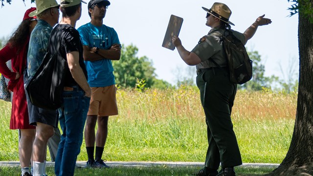 A uniformed park ranger speaks to a group under a tree