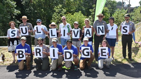A group of volunteers, youth, and park rangers hold up letters to spell 