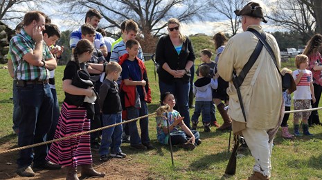a ranger dressed like an 18th century soldier speaks to adults and children. some kids are seated.