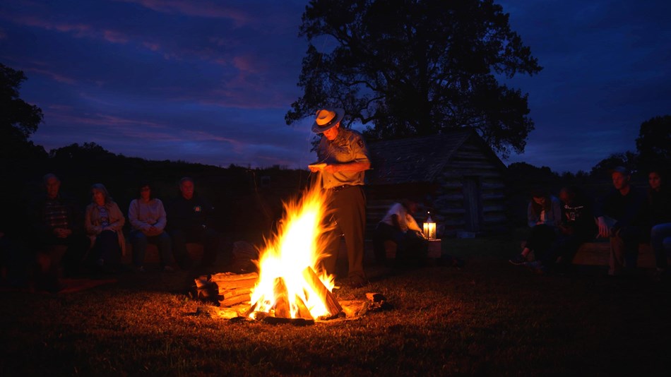a ranger stands next to a glowing campfire after dark and people sit behind on benches