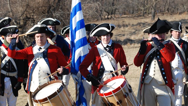 Continental Army musicians stand in line in front of a regiment outdoors in a field.