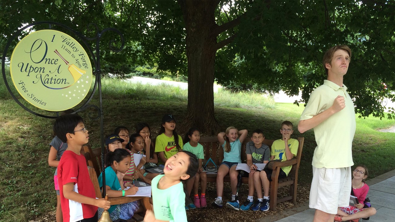 A young story teller entertains a group of children at an outdoor bench.