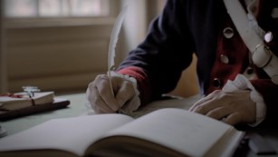 an open book sits on a desk and a hand writes with a quill pen