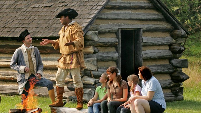 A couple of men in colonial clothing tell stories to a group of children near some log huts.