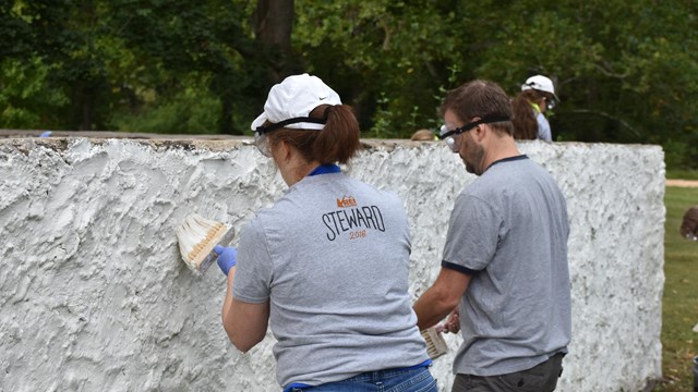 Two adults holding paint brushes whitewash a wall.
