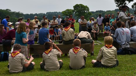 Children in Boy Scout uniforms sit in the grass watching a ranger program.