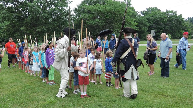 Soldiers teaching a group of children how to drill with wooden muskets. 