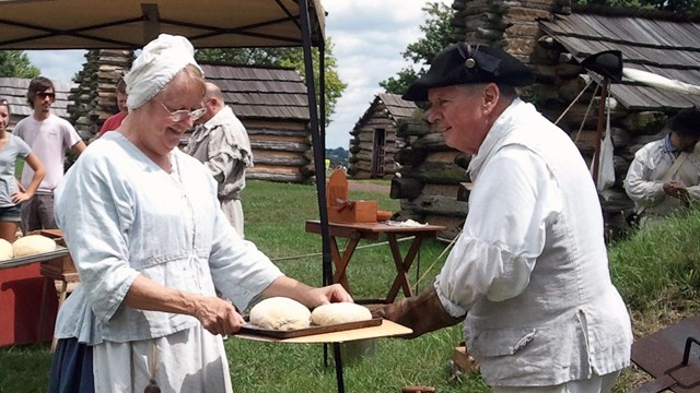 Two colonial women bring bread dough to a colonial baker at an outdoor oven.