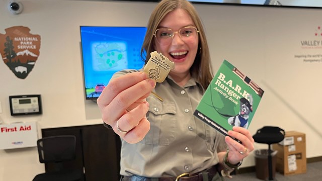 A smiling park ranger holds up a dog tag and activity book in the visitor center.