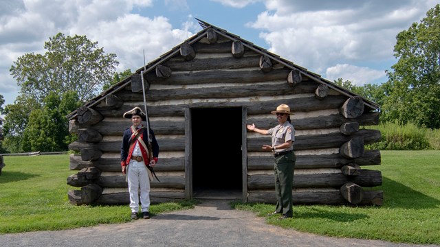 two rangers, one in a soldier uniform and another in the NPS uniform stand outside a hut