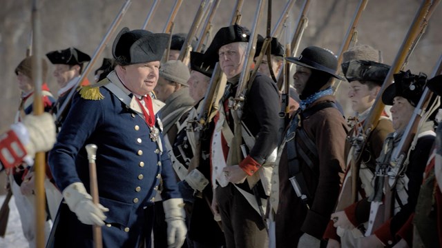Soldiers holding muskets stand at attention in front of Baron von Steuben