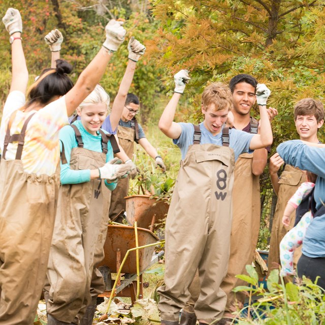 A grooup of kids wearing gloves and coveralls smile with hands in the air