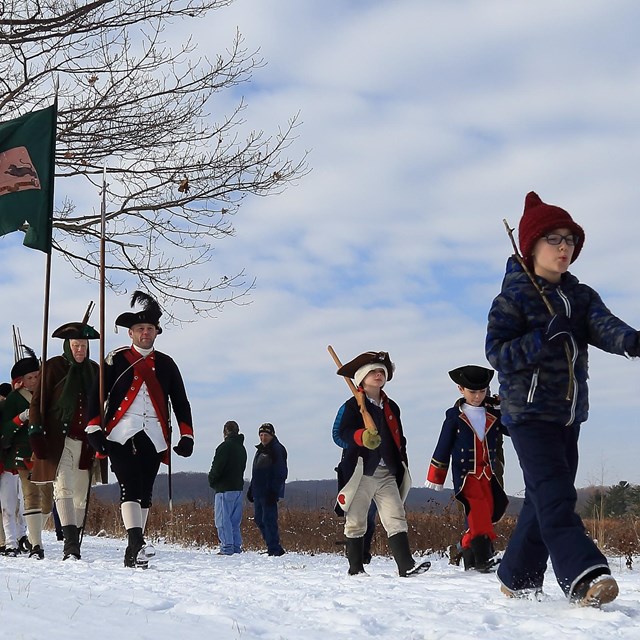 children march in front of reenactors wearing the clothing of 18th century soldiers