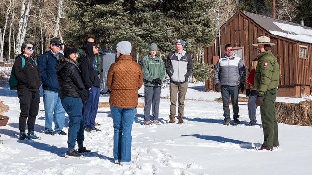 A group of people gather around a park ranger in the snow near a historic cabin.