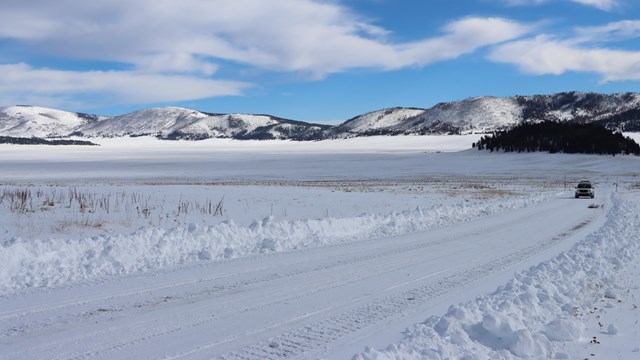 A car drives up a snow-covered road winding through a vast, snowy grassland.