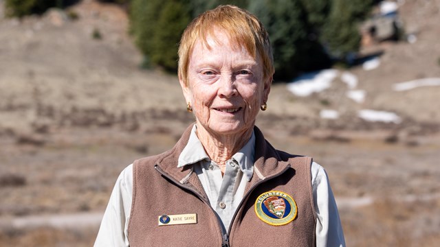 A park volunteer wearing a brown uniform vest over a khaki shirt smiles at us.
