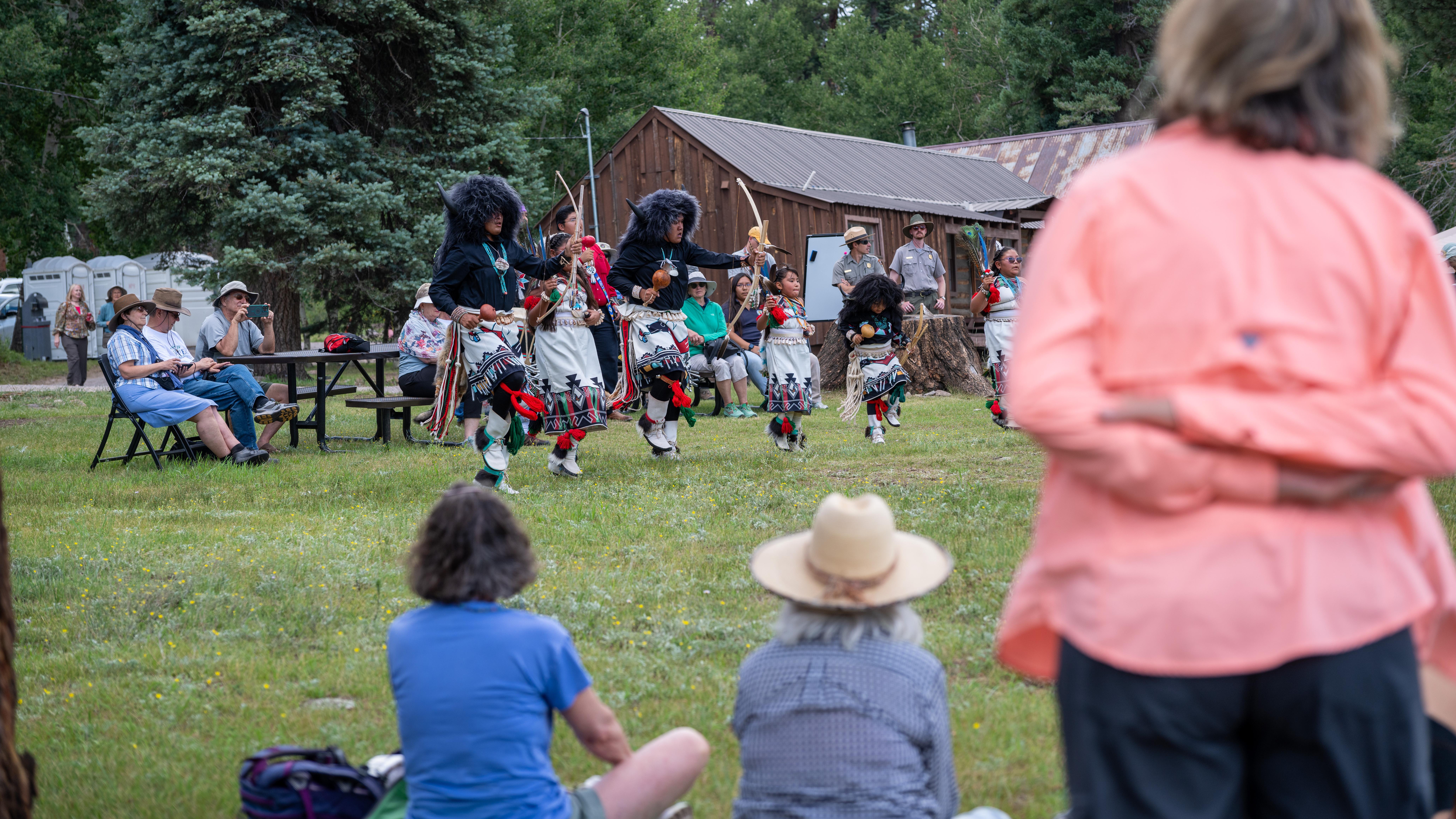A pueblo dance group performs outdoors in front of an audience.