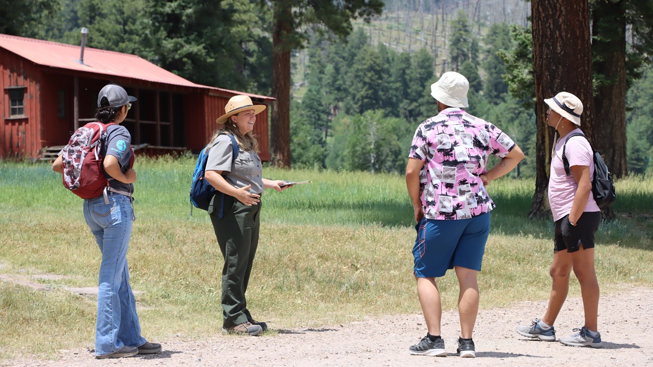 A park ranger addresses 3 visitors while standing in front of a historic cabin.