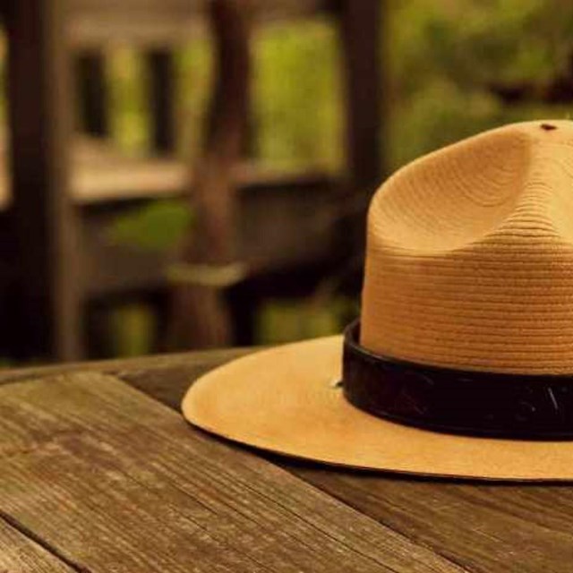 A National Park Service ranger hat rests on a wooden picnic table in the woods.